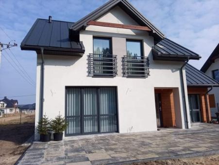 Nowoczesny, dwukondygnacyjny dom z białymi ścianami, ciemną dachówką, szarymi ramami okiennymi i balkonami z metalowymi balustradami, otoczony kostką brukową i młodymi drzewkami iglastymi w donicach.