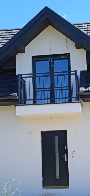 Nowoczesny balkon z czarną metalową balustradą na tle białej fasady domu z ciemnym dachem, widoczna również czarna brama wejściowa z przeszkleniem.