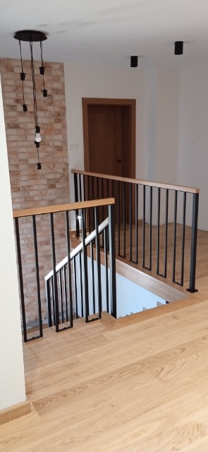 Jasne drewniane schody z czarną metalową balustradą i drewnianym poręczą, widok z góry, ściana z cegły w tle, nowoczesne oświetlenie sufitowe.