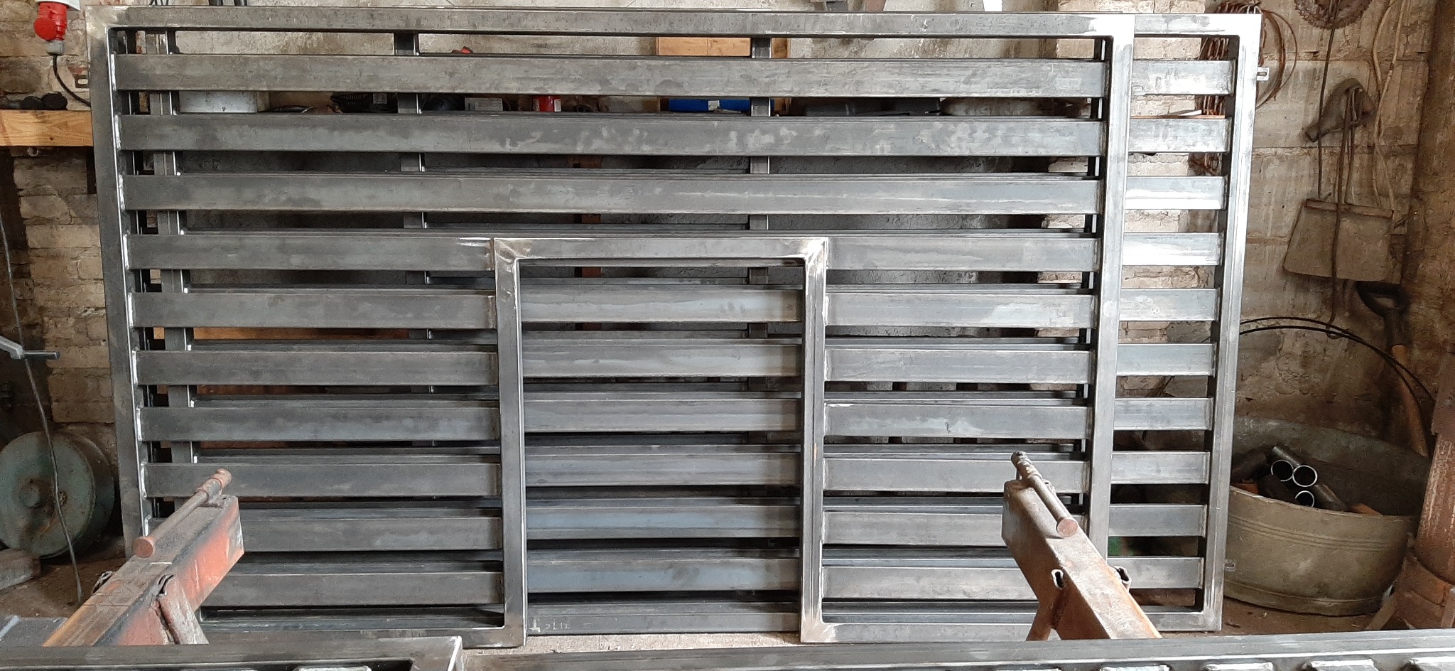 Surowy, spawany element metalowy ogrodzenia z poziomymi szczeblami i ramą, w warsztacie z narzędziami i ceglaną ścianą w tle.