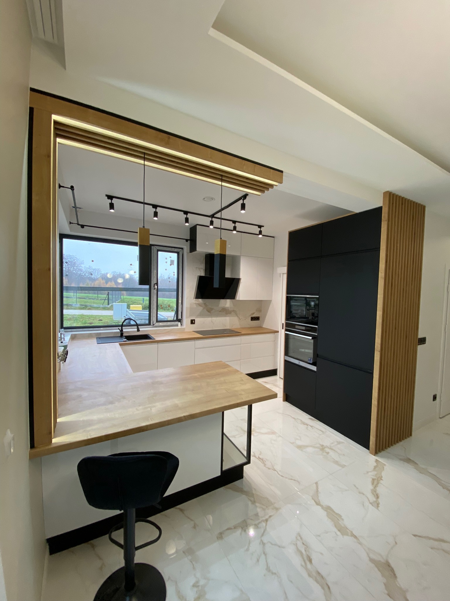 Nowoczesna kuchnia z wyspą, białe fronty szafek, czarny słupek z piekarnikiem i mikrofalówką, drewniane akcenty, oświetlenie szynowe i marmurowa podłoga, widok na ogród za oknem.