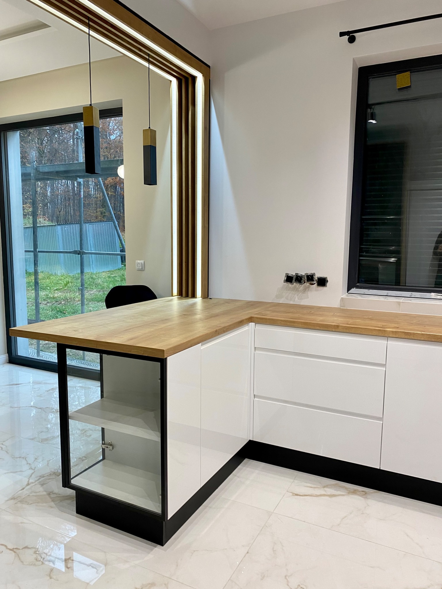 Kąt kuchni z białymi szafkami, drewnianym blatem i czarnymi akcentami, widok na okno z ogrodem i niedokończoną konstrukcją.