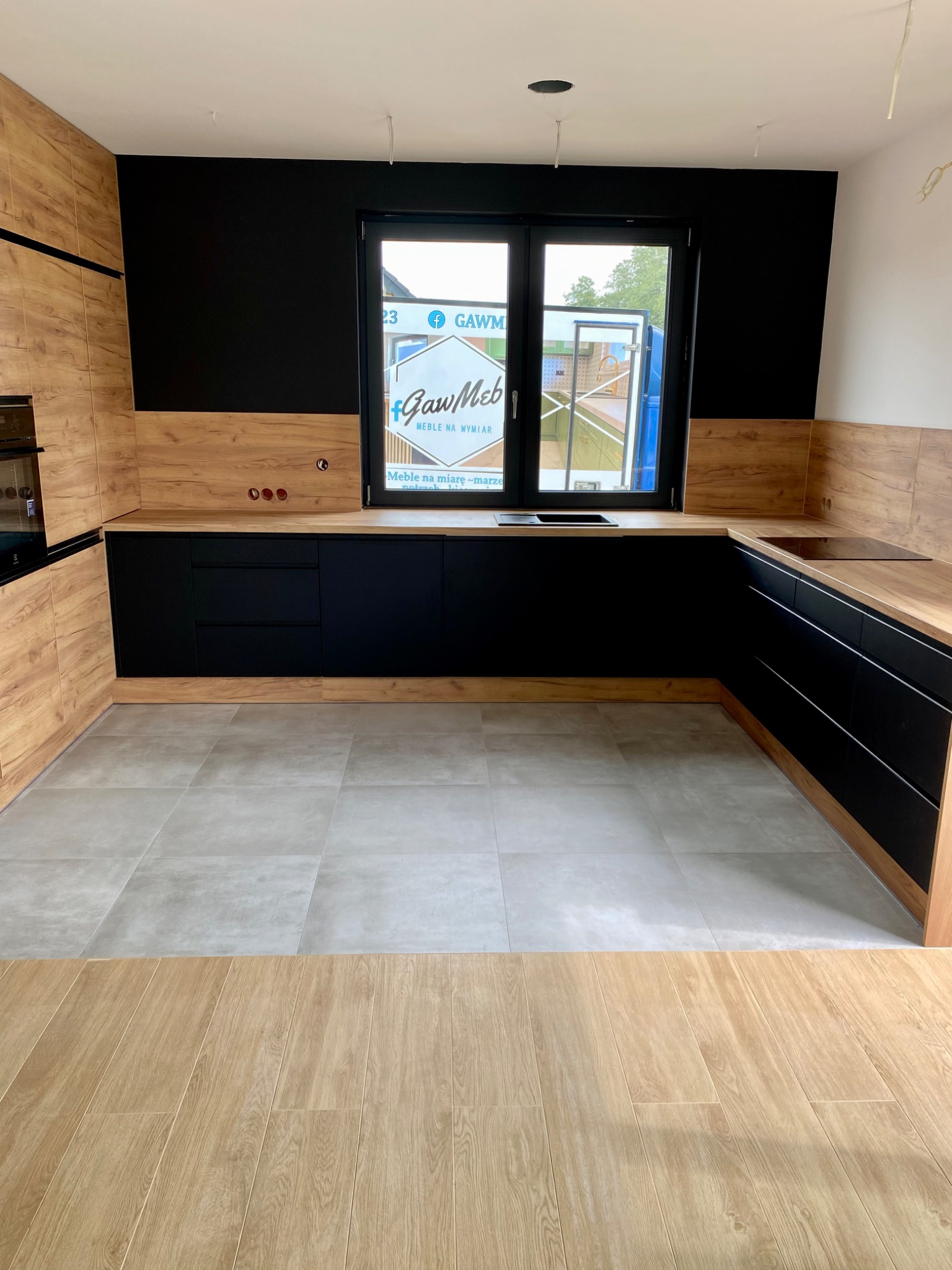 Nowoczesna kuchnia z czarnymi frontami i drewnopodobnymi blatami, szara podłoga, okno z widocznym logotypem firmy meblarskiej, surowy stan wykończenia.