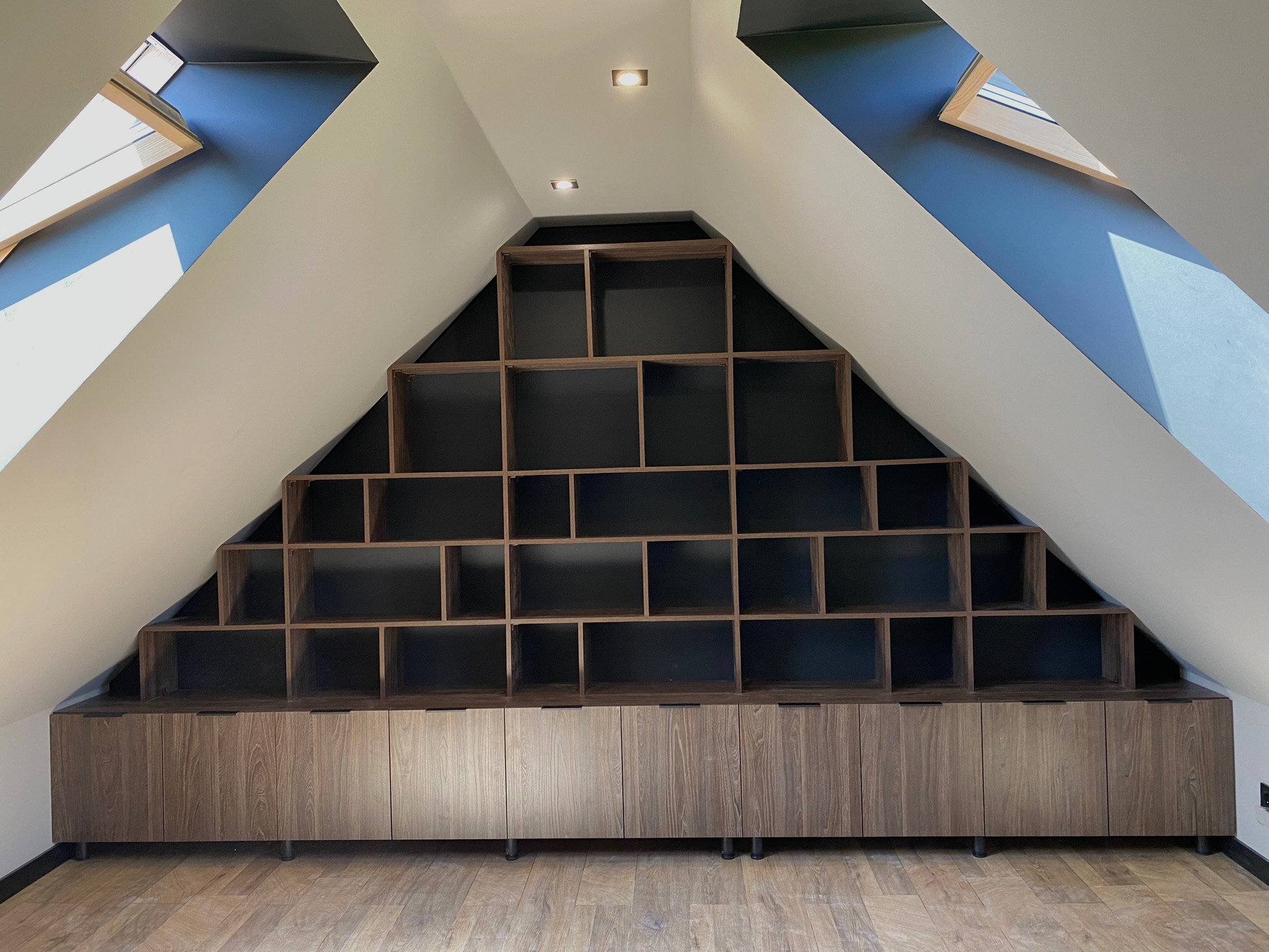 Szafa wnękowa z drewnianymi frontami i otwartymi półkami w kształcie schodów, dopasowana do skosu poddasza, oświetlona punktowymi lampami sufitowymi