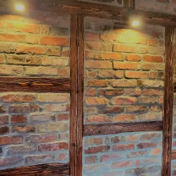 Dekoracyjna ściana z czerwonej cegły z drewnianymi belkami, oświetlona trzema punktowymi lampami sufitowymi.