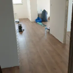 Wnętrze w trakcie remontu, widoczna nowa, brązowa podłoga laminowana, białe ściany, surowe wykończenie ościeżnicy drzwiowej, worki z gruzem i narzędzia w tle.