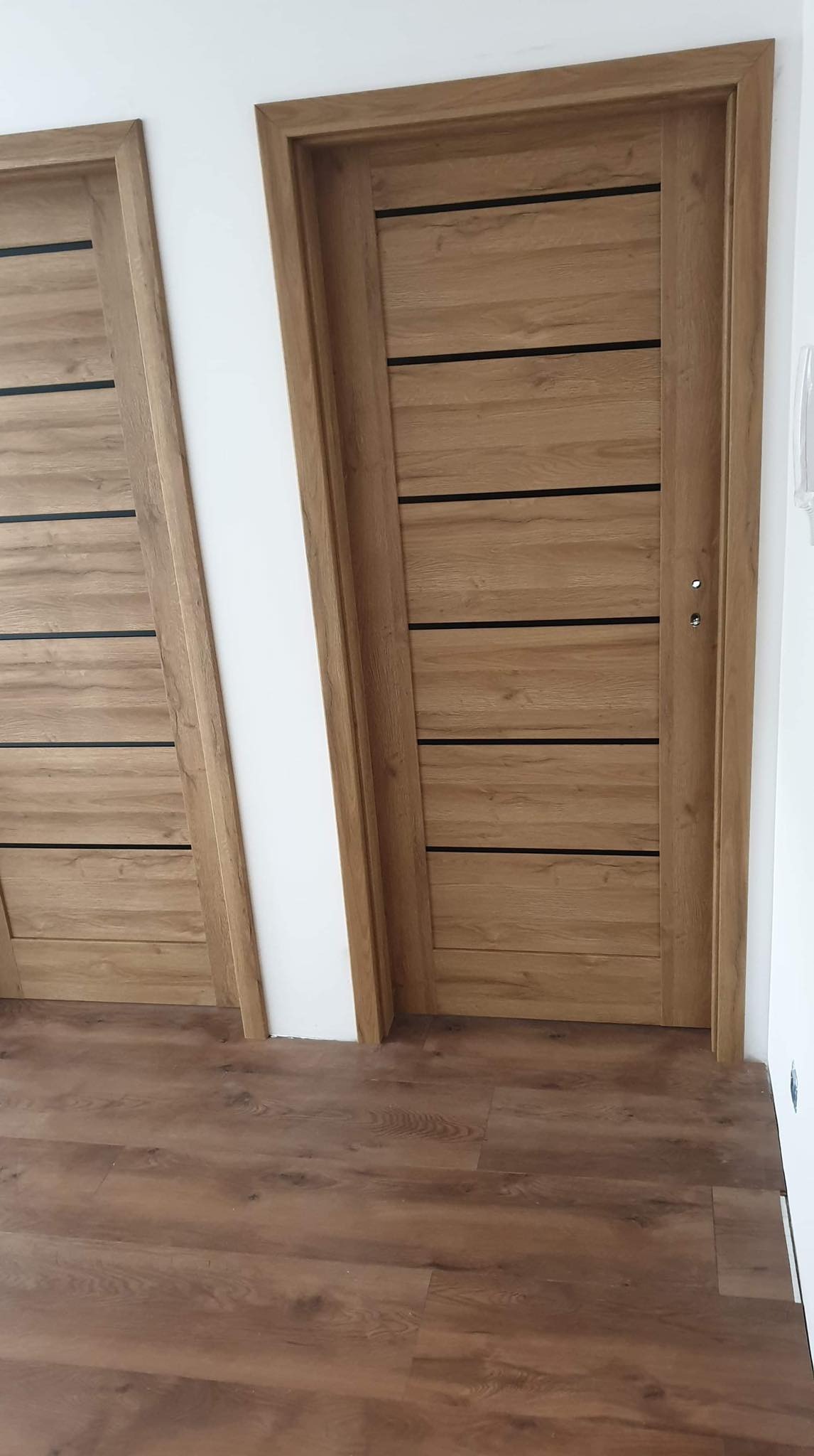 Dwa nowe, drewniane drzwi wewnętrzne z poziomymi, czarnymi wstawkami, zamontowane w białej ścianie na podłodze z paneli drewnopodobnych.