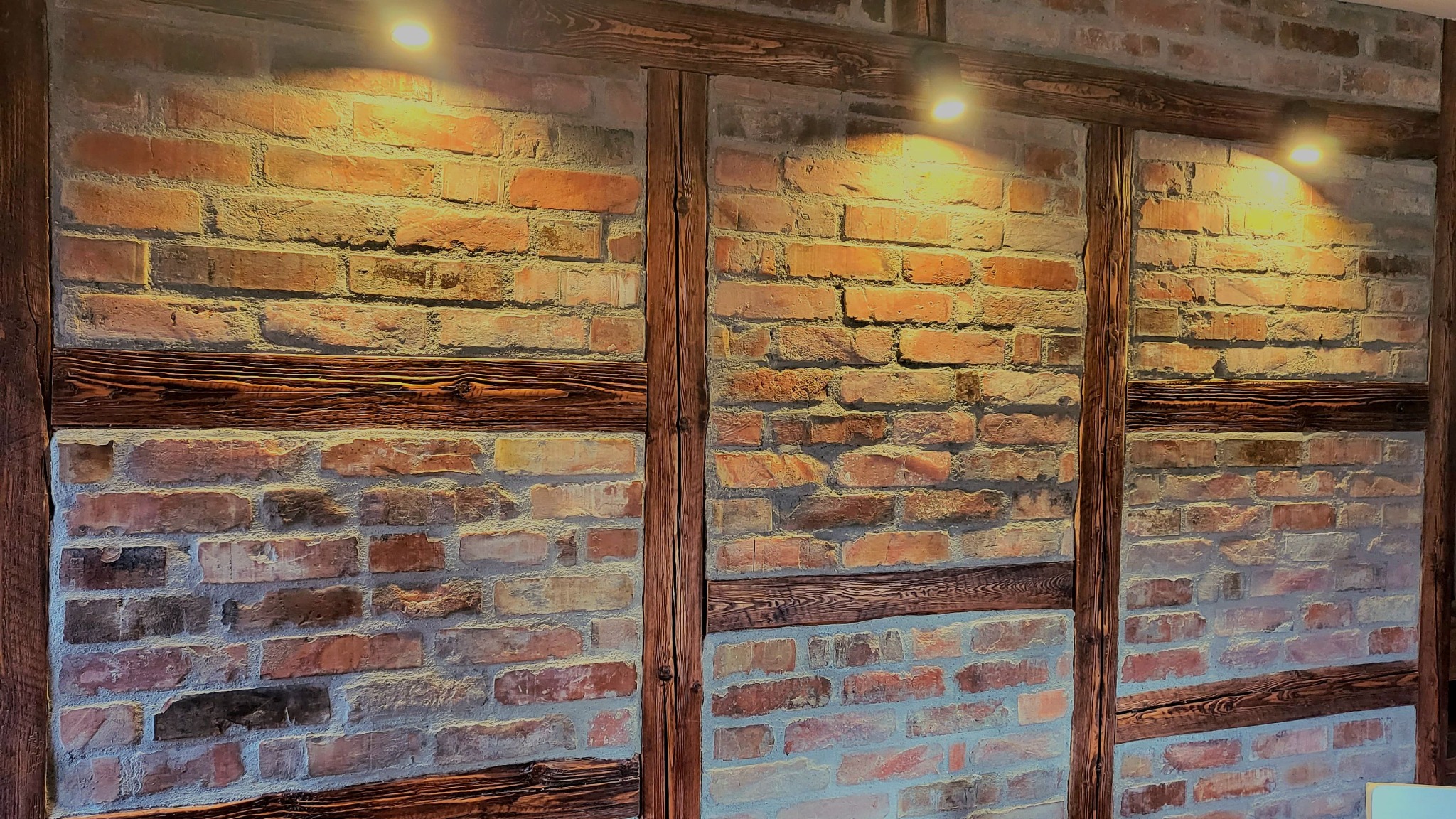 Dekoracyjna ściana z czerwonej cegły z drewnianymi belkami, oświetlona trzema punktowymi lampami sufitowymi.