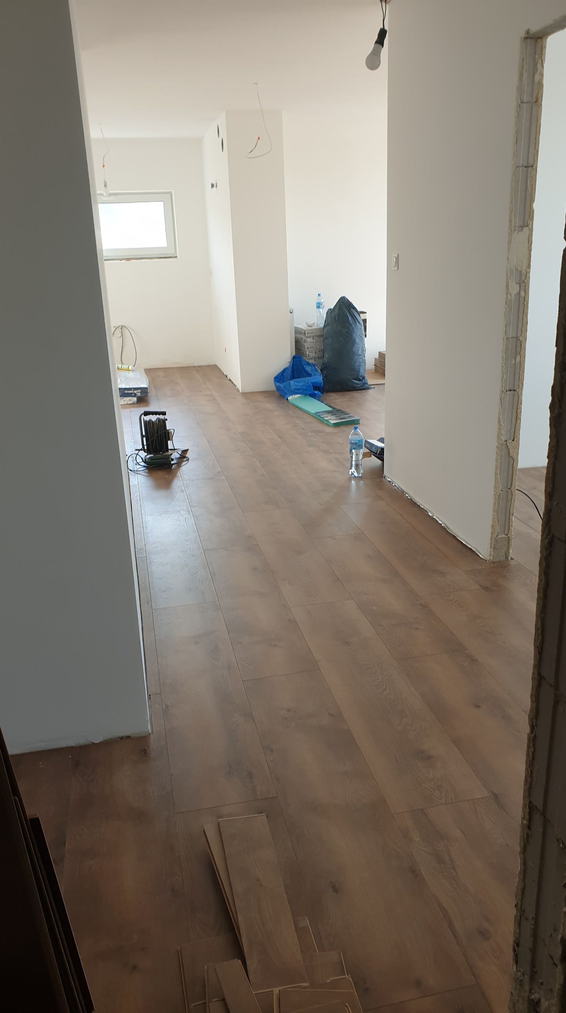 Wnętrze w trakcie remontu, widoczna nowa, brązowa podłoga laminowana, białe ściany, surowe wykończenie ościeżnicy drzwiowej, worki z gruzem i narzędzia w tle.