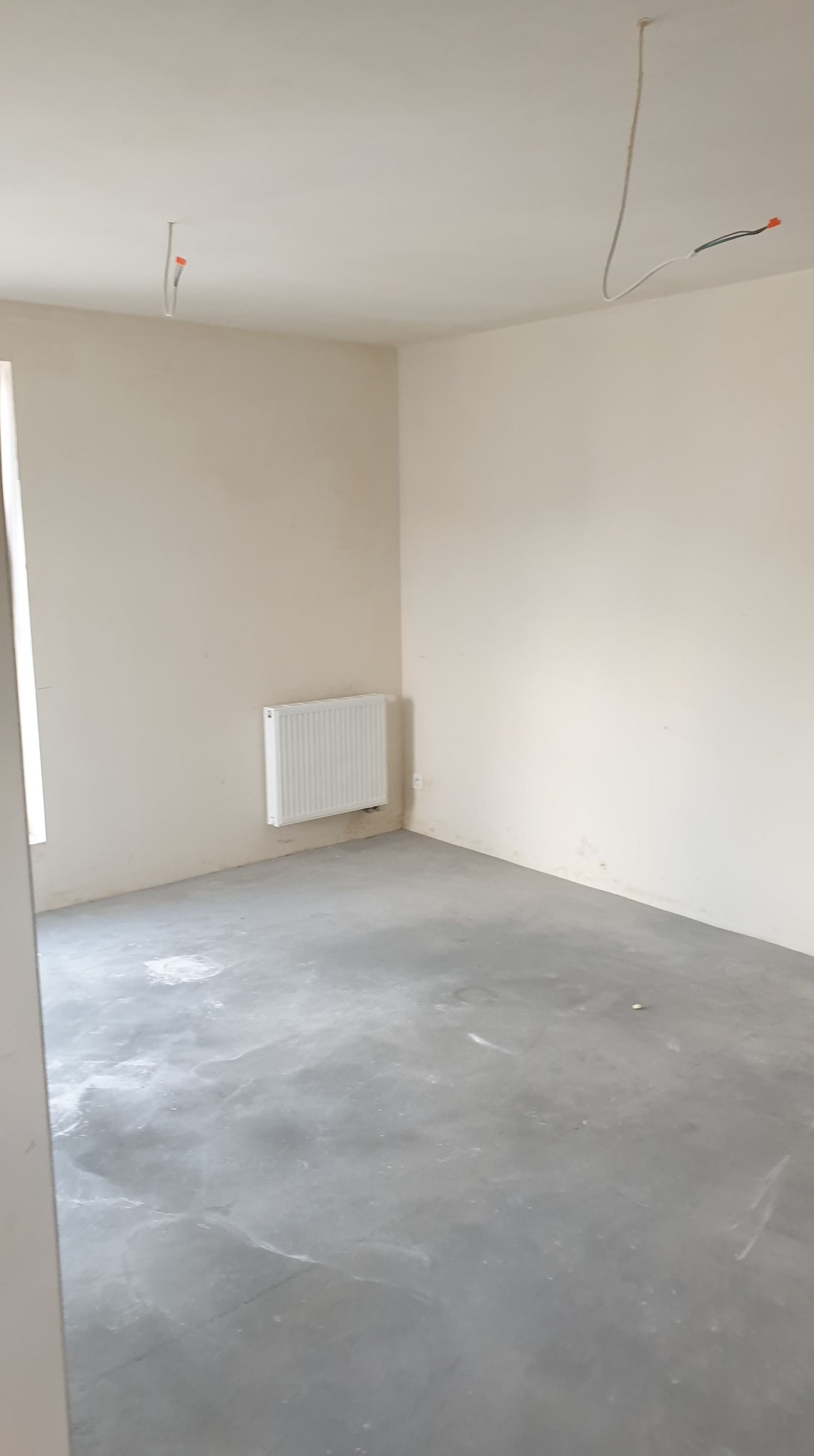 Pusty pokój w stanie deweloperskim z surową podłogą, świeżo pomalowanymi ścianami w odcieniach beżu, białym grzejnikiem na ścianie i wystającymi kablami z sufitu.