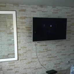 Nowe okno PCV z białymi ramami w pokoju z dekoracyjną ścianą imitującą kamień, na której wisi telewizor.