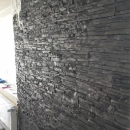 Ściana wykończona ciemnym kamieniem dekoracyjnym, widoczne gniazdko elektryczne, częściowo zakryte okno z zabezpieczeniem folią malarską.