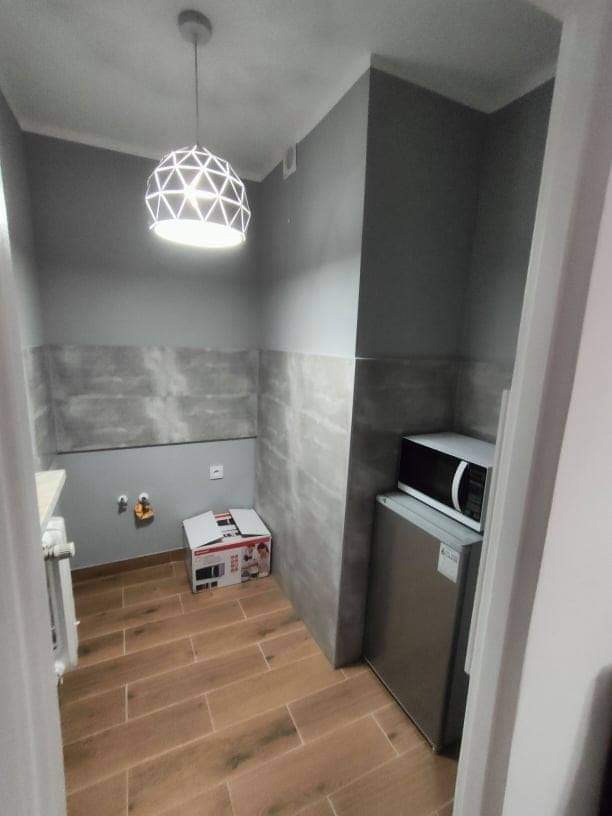 Mała kuchnia po remoncie z drewnopodobnymi panelami na podłodze i szarymi płytkami na ścianach, widoczna lampa sufitowa, lodówka i mikrofalówka, karton po mikrofalówce leży na podłodze.