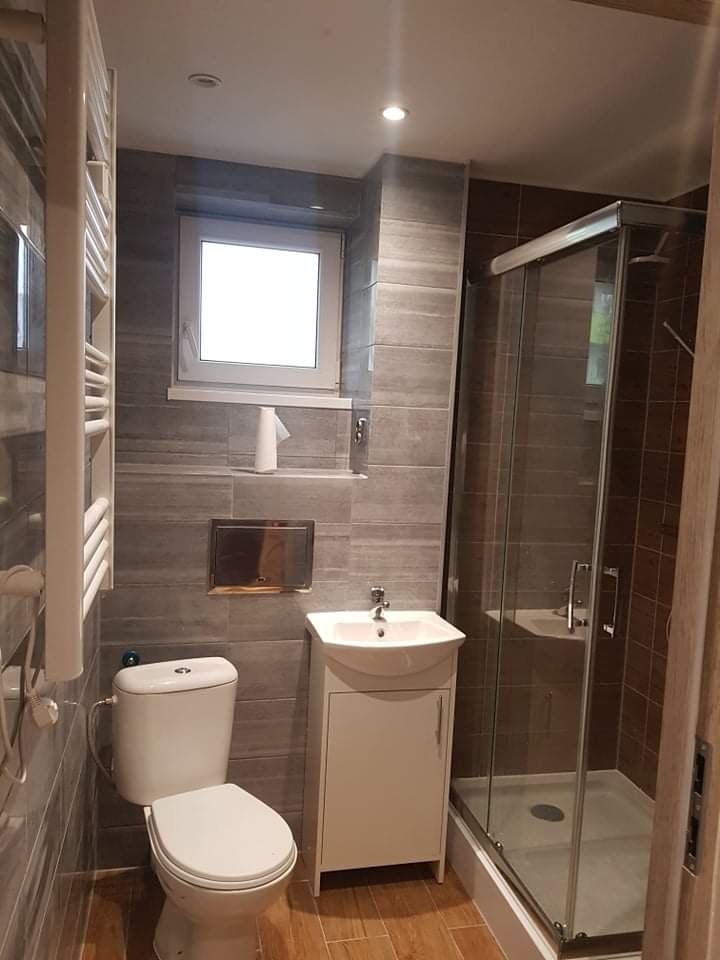 Nowoczesna łazienka z szaro-brązowymi płytkami imitującymi drewno, kabiną prysznicową z przezroczystego szkła, białą umywalką z szafką, toaletą i grzejnikiem drabinkowym. Okno z białymi ramami...