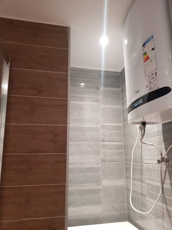 Wykończona łazienka z brązowymi i szarymi płytkami imitującymi drewno, widoczny biały bojler elektryczny Tesy zamontowany na ścianie.