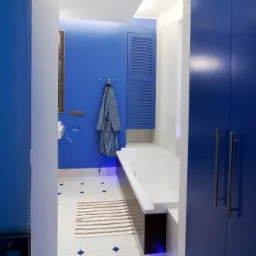 Niebieska łazienka z wanną, grzejnikiem żeberkowym i szlafrokiem w paski wiszącym na haczyku, widok zza uchylonych drzwi.