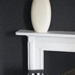 Kremowy wazon na białym kominku z narzędziami kominkowymi na tle ciemnej ściany.