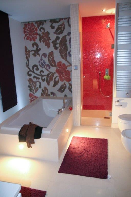 Nowoczesna łazienka z białą wanną obudowaną płytkami, kabiną prysznicową z czerwoną mozaiką i wzorzystą ścianą z motywem kwiatowym, uzupełniona burgundowym dywanikiem.
