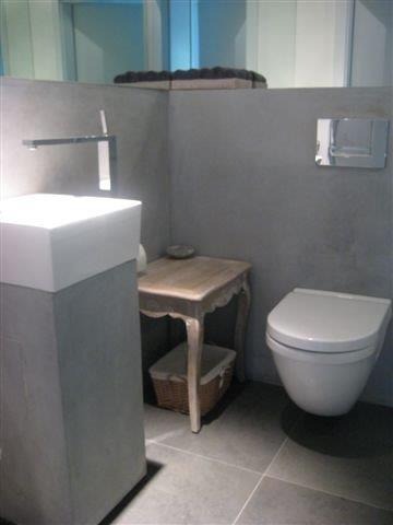 Nowoczesna łazienka z szarą, betonową ścianą, wiszącą toaletą, umywalką na postumencie i małym drewnianym stolikiem z koszem.