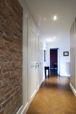 Długi korytarz z parkietem ułożonym we wzór jodełki, ceglana ściana z widoczną fakturą, białe szafy w zabudowie i wąski stolik na końcu korytarza.