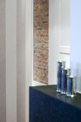 Fragment wnętrza z tapetą w pionowe pasy, ceglaną ścianą w głębi i metalowymi dekoracjami na ciemnym blacie.