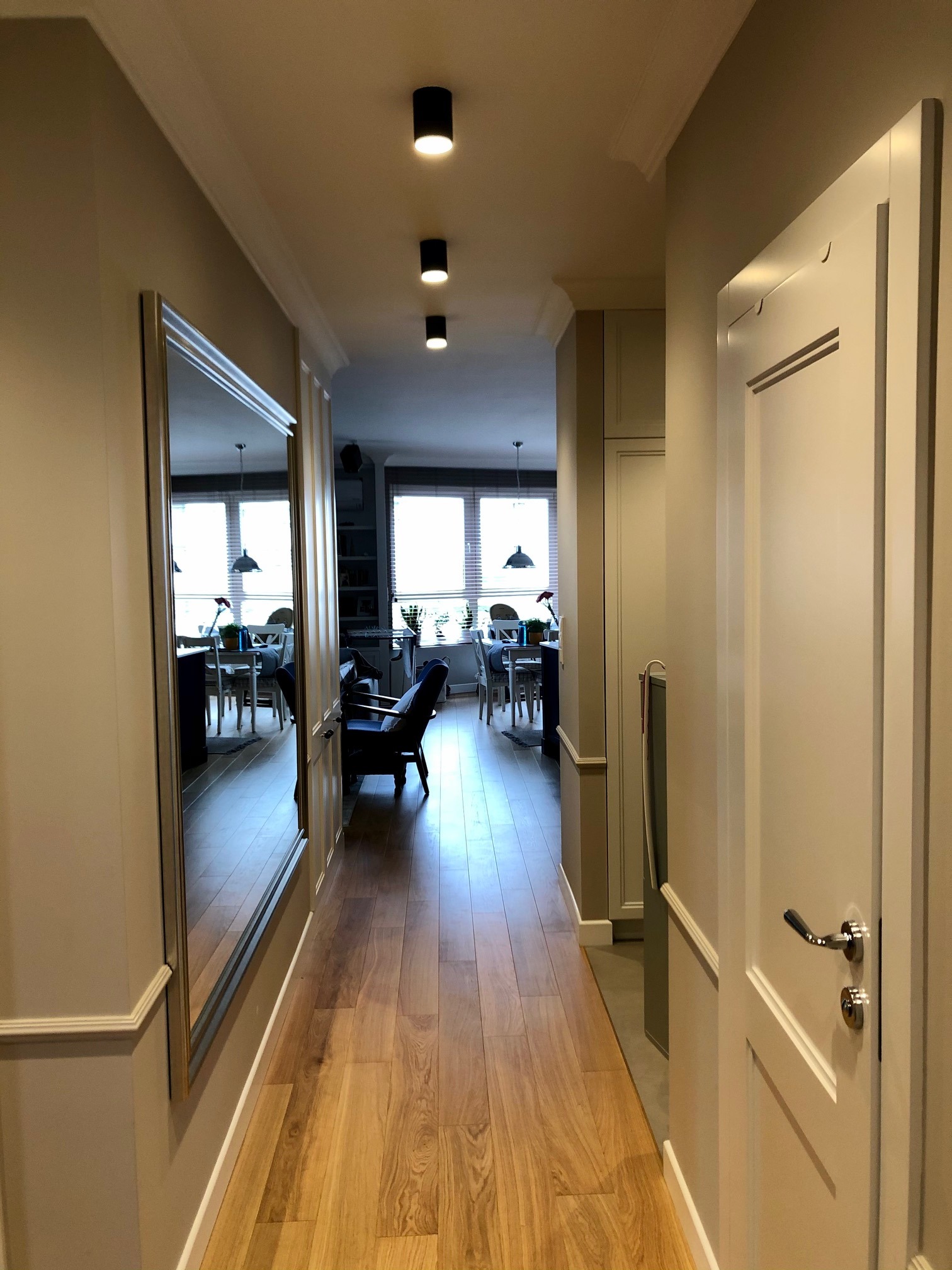 Korytarz z drewnianą podłogą, lustrem w złotej ramie i widokiem na salon z niebieskim fotelem i jadalnią.