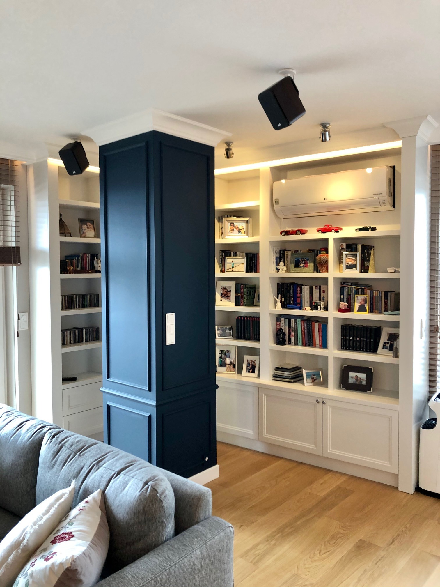 Salon z zabudową biblioteczną: białe półki z książkami i dekoracjami, klimatyzator na górze, ciemnoniebieska kolumna z ukrytymi drzwiami, głośniki na suficie, szara sofa z poduszkami na pierwszym...