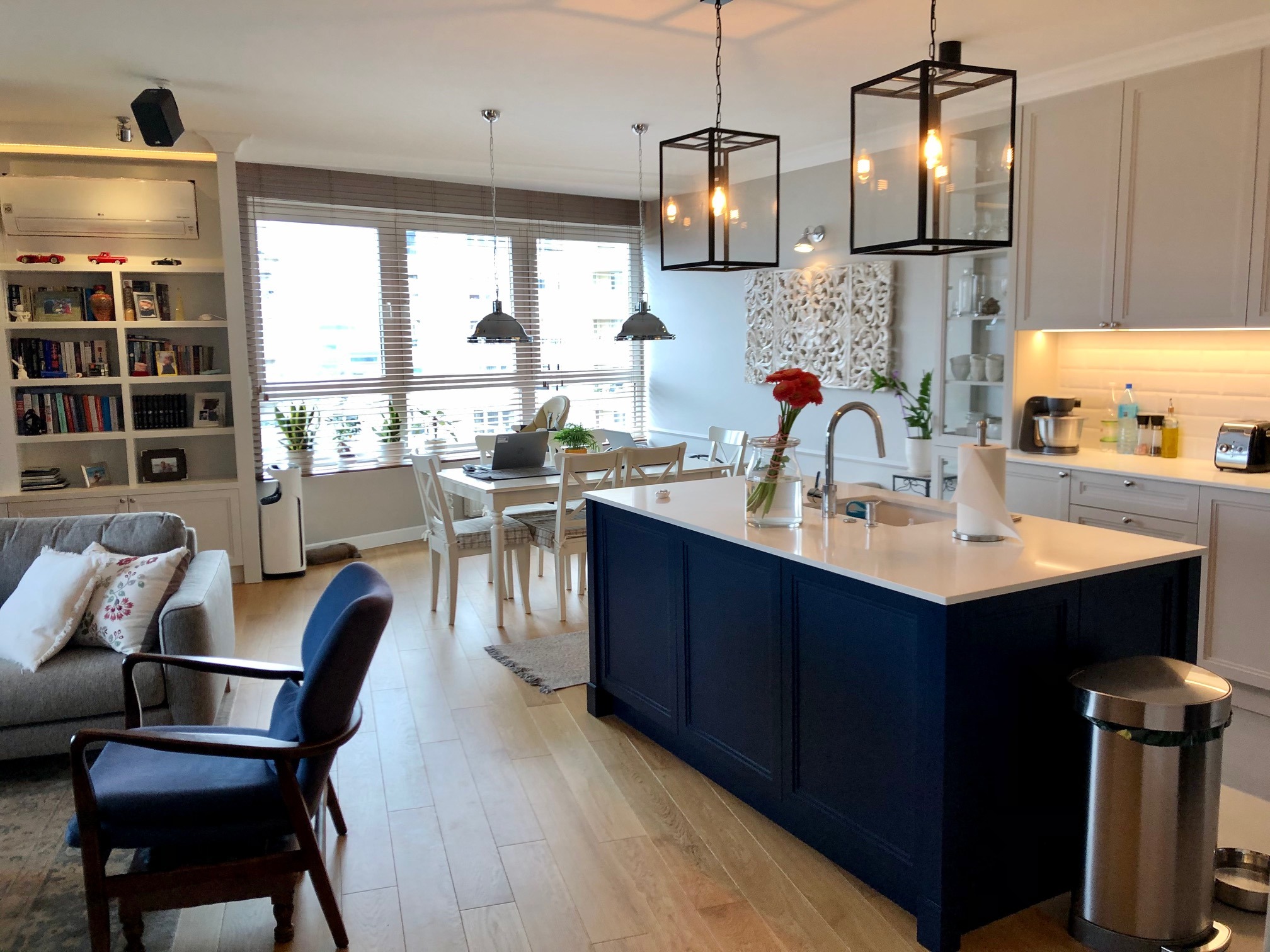 Jasne wnętrze z drewnianą podłogą, wyspą kuchenną w kolorze granatowym, białymi szafkami i stołem jadalnym z laptopem przy oknie.
