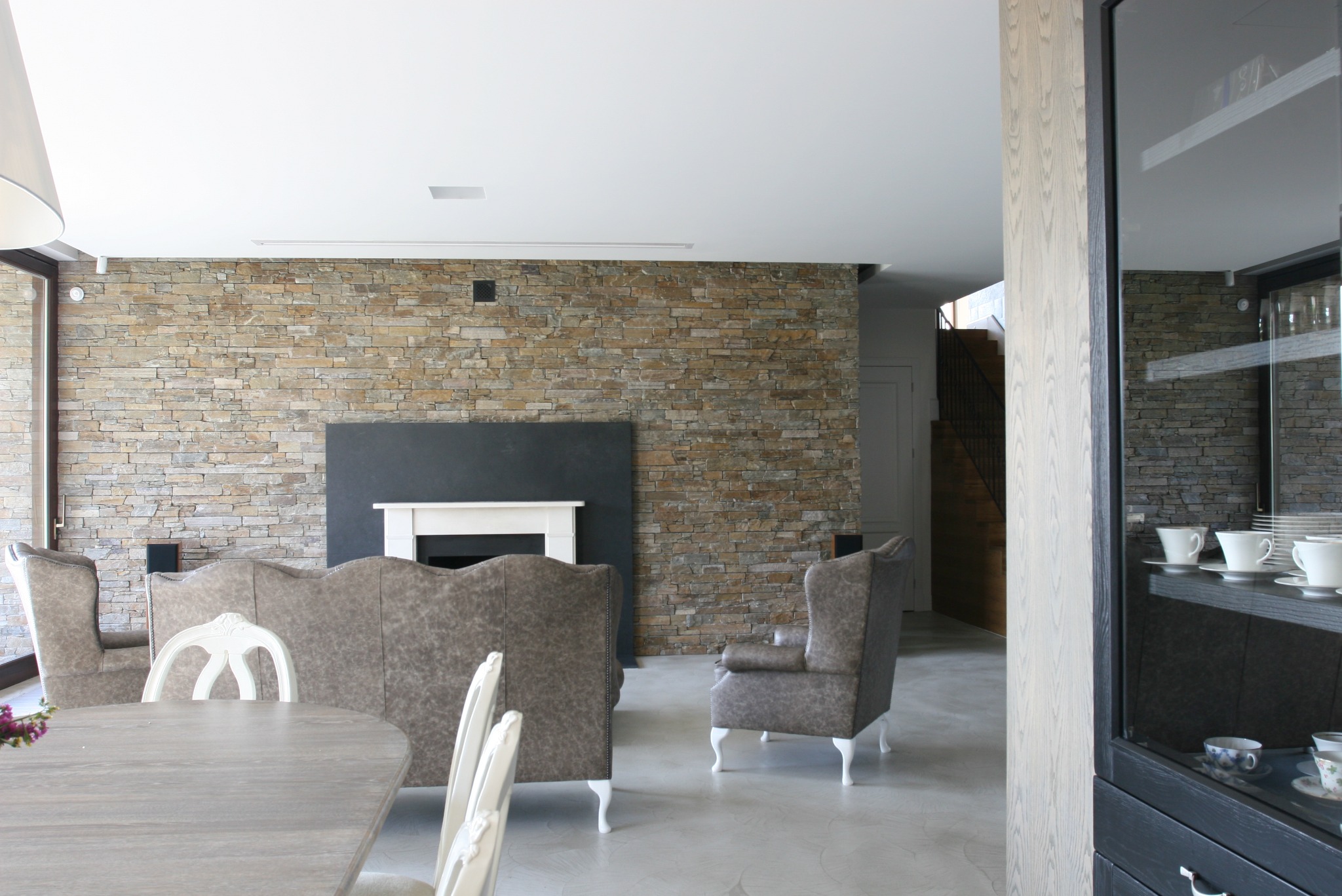 Salon z kamienną ścianą, kominkiem, szarą sofą i fotelami w stylu vintage, widok na schody i przeszkloną witrynę z porcelaną.