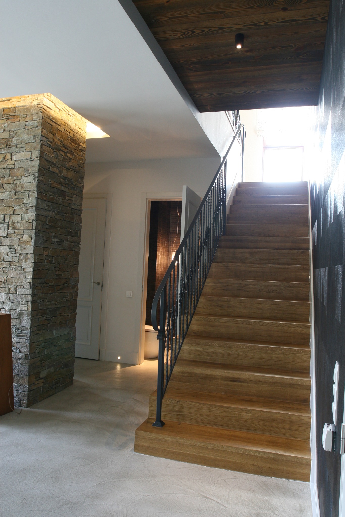 Wnętrze domu z drewnianymi schodami z czarną balustradą, kamienną kolumną i drewnianym sufitem z oświetleniem punktowym.