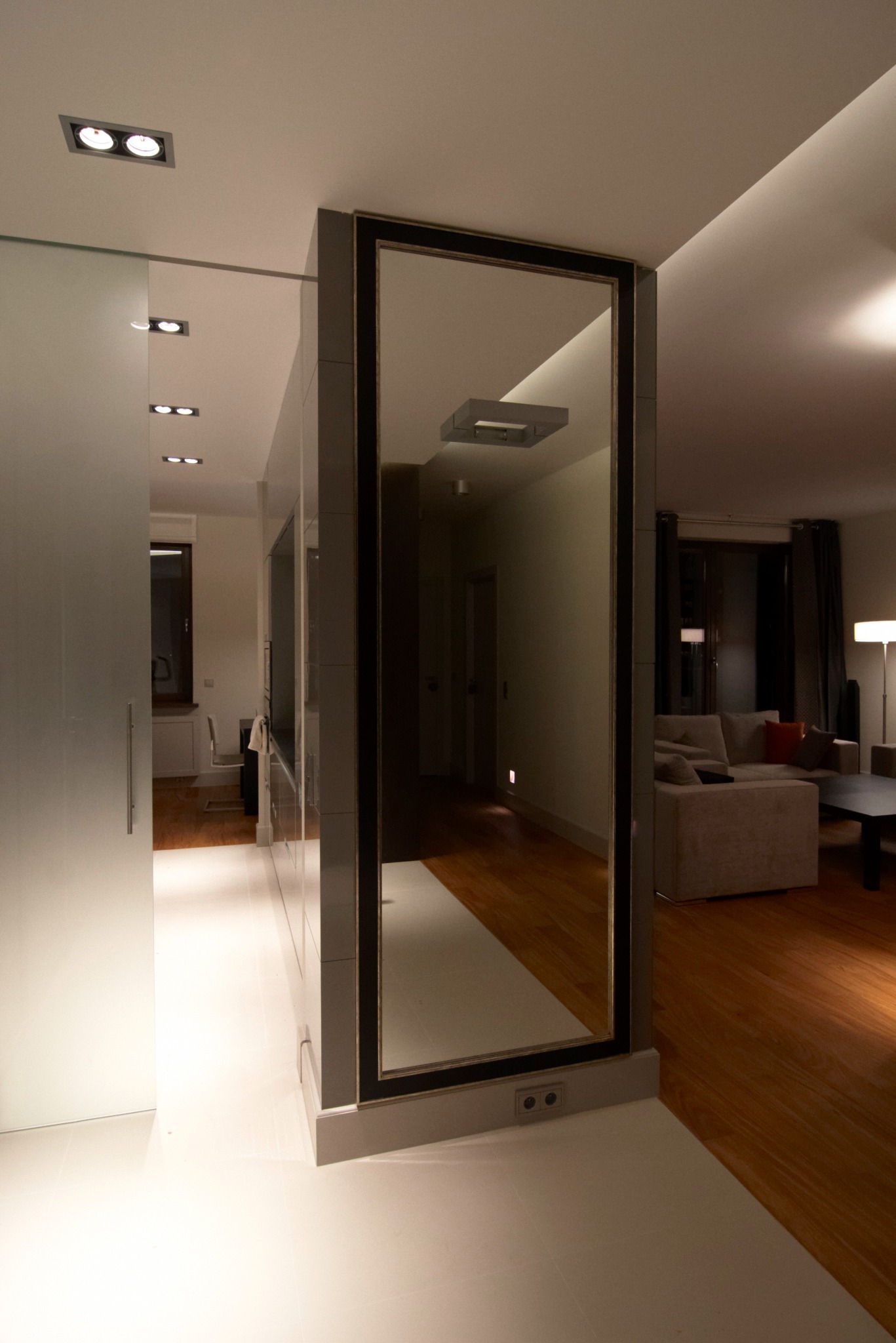 Nowoczesne wnętrze z dużym lustrem w szarej ramie, oddzielającym białą kuchnię od salonu z drewnianą podłogą i beżową sofą.