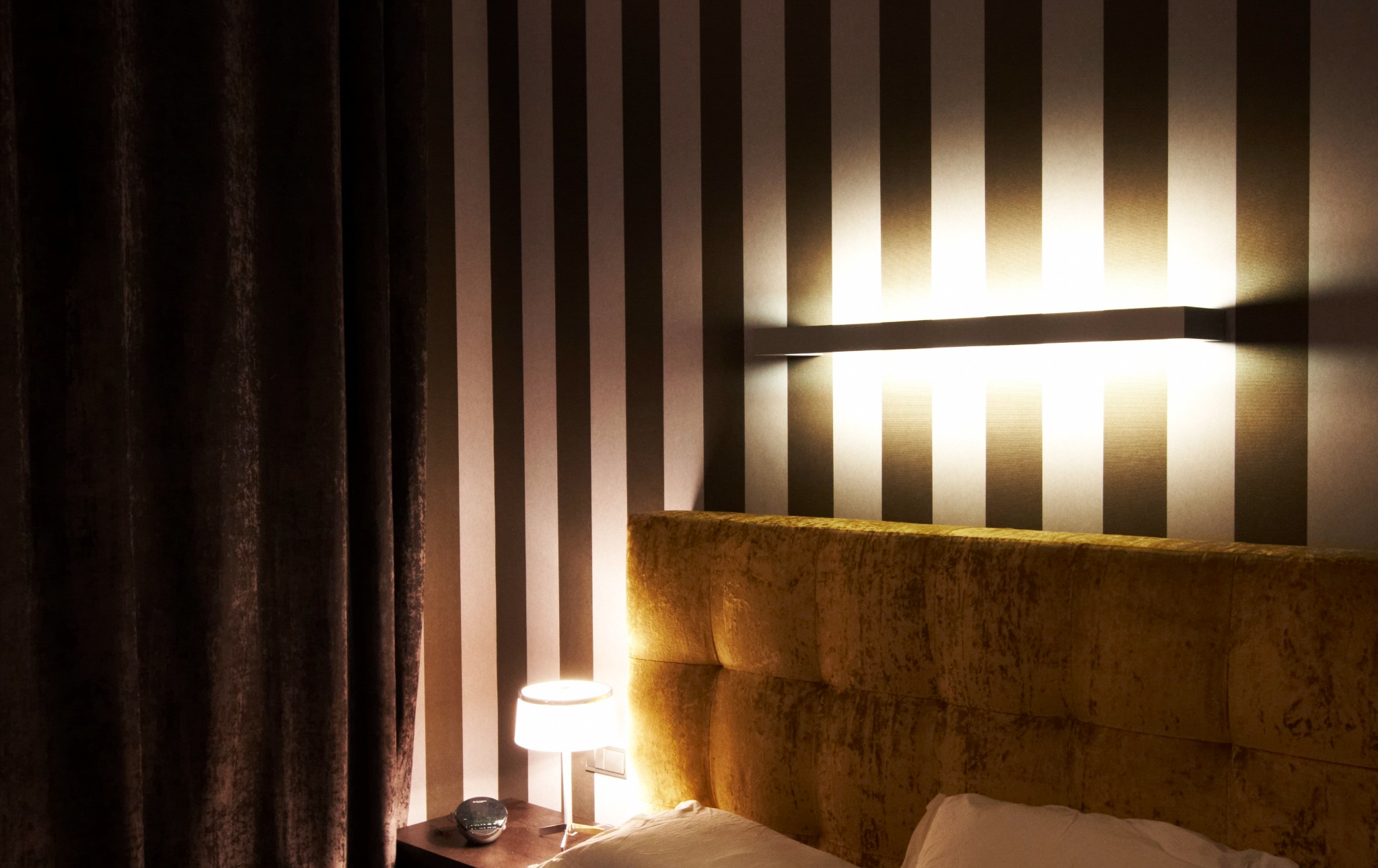 Sypialnia z tapetą w pionowe pasy, podświetlona listwą LED nad wezgłowiem łóżka, obok zasłonięte okno i lampka nocna na stoliku.