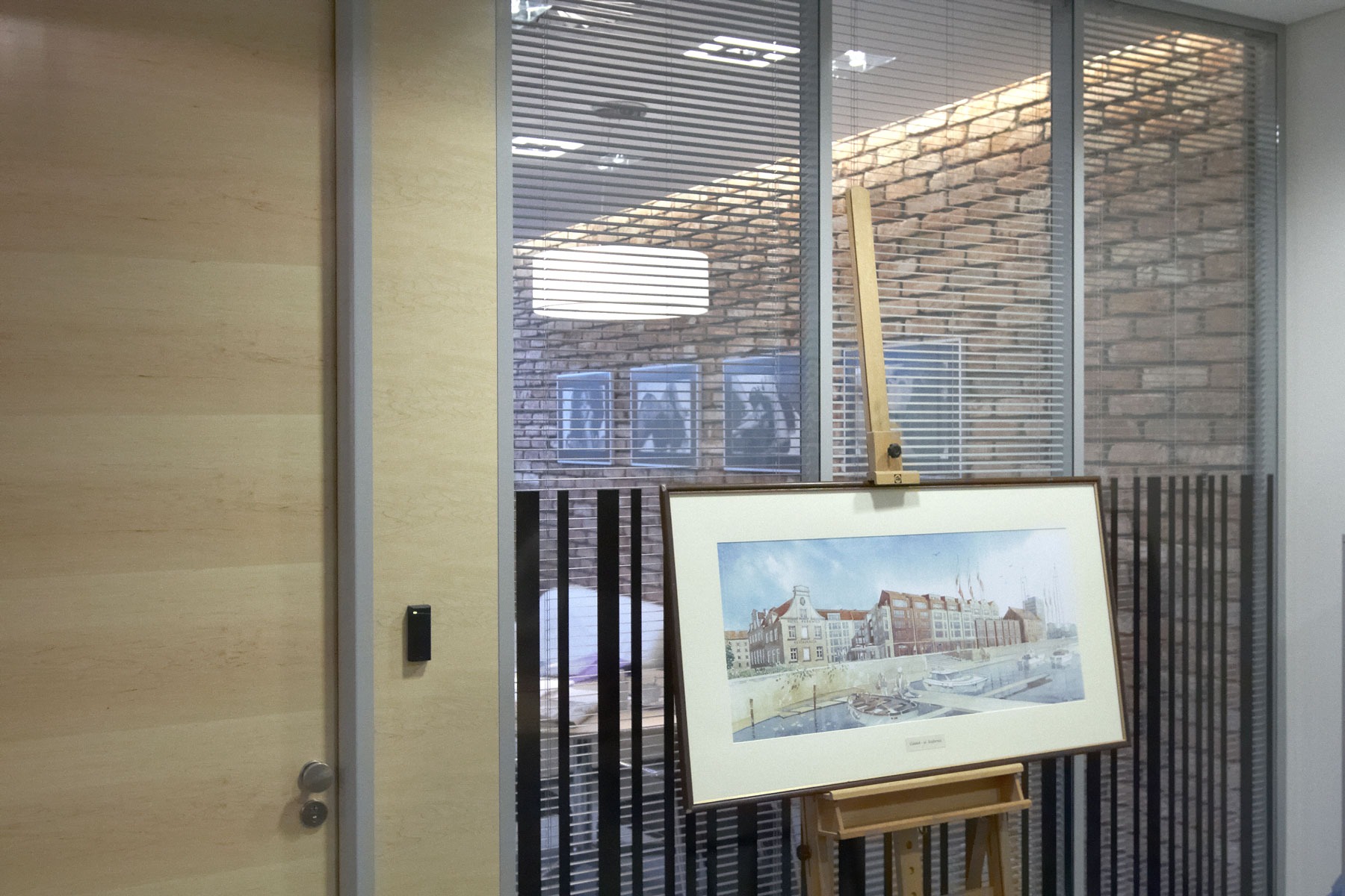 Obraz z motywem portowego miasteczka na sztaludze w biurze, widok przez żaluzje na ceglaną ścianę i powieszone grafiki.