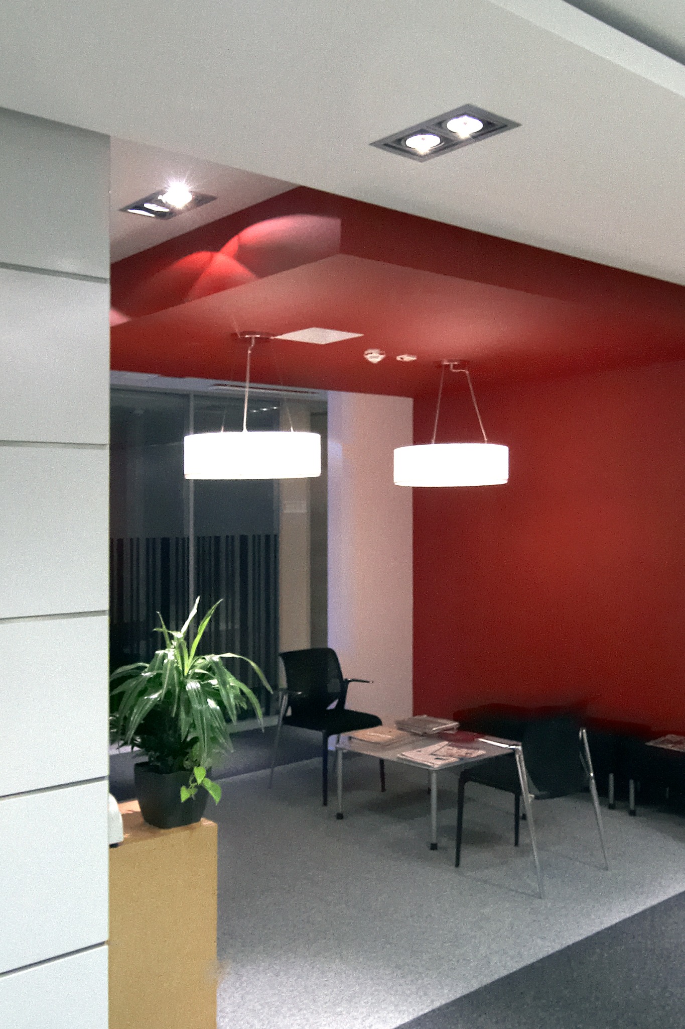 Nowoczesna poczekalnia biurowa z czerwonym sufitem, okrągłymi lampami wiszącymi, czarnymi krzesłami i stolikiem z broszurami.