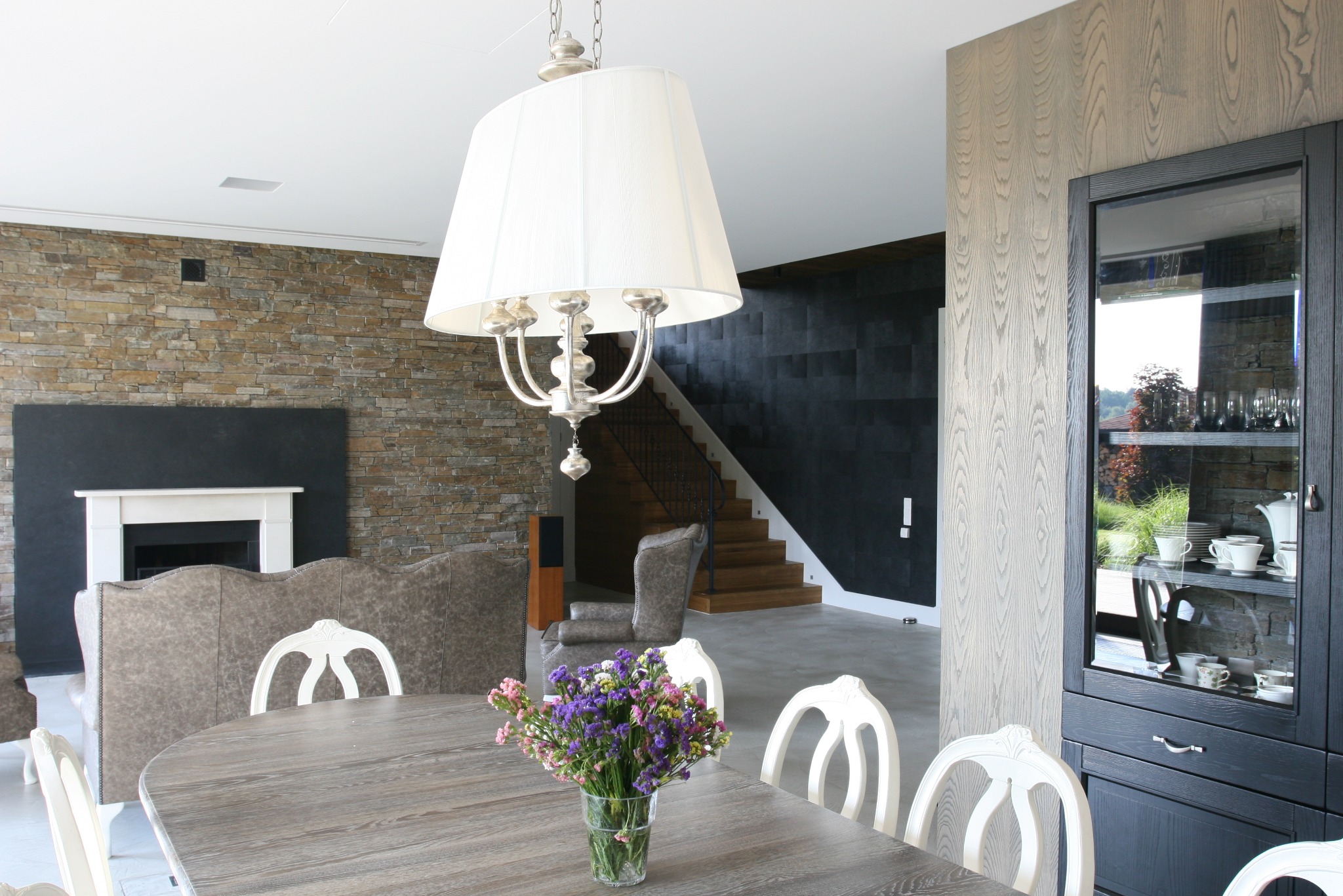 Jasne, przestronne wnętrze jadalni w Warszawie z drewnianym stołem, białymi krzesłami, kwiatami w wazonie, widokiem na schody i kamienną ścianę z kominkiem.
