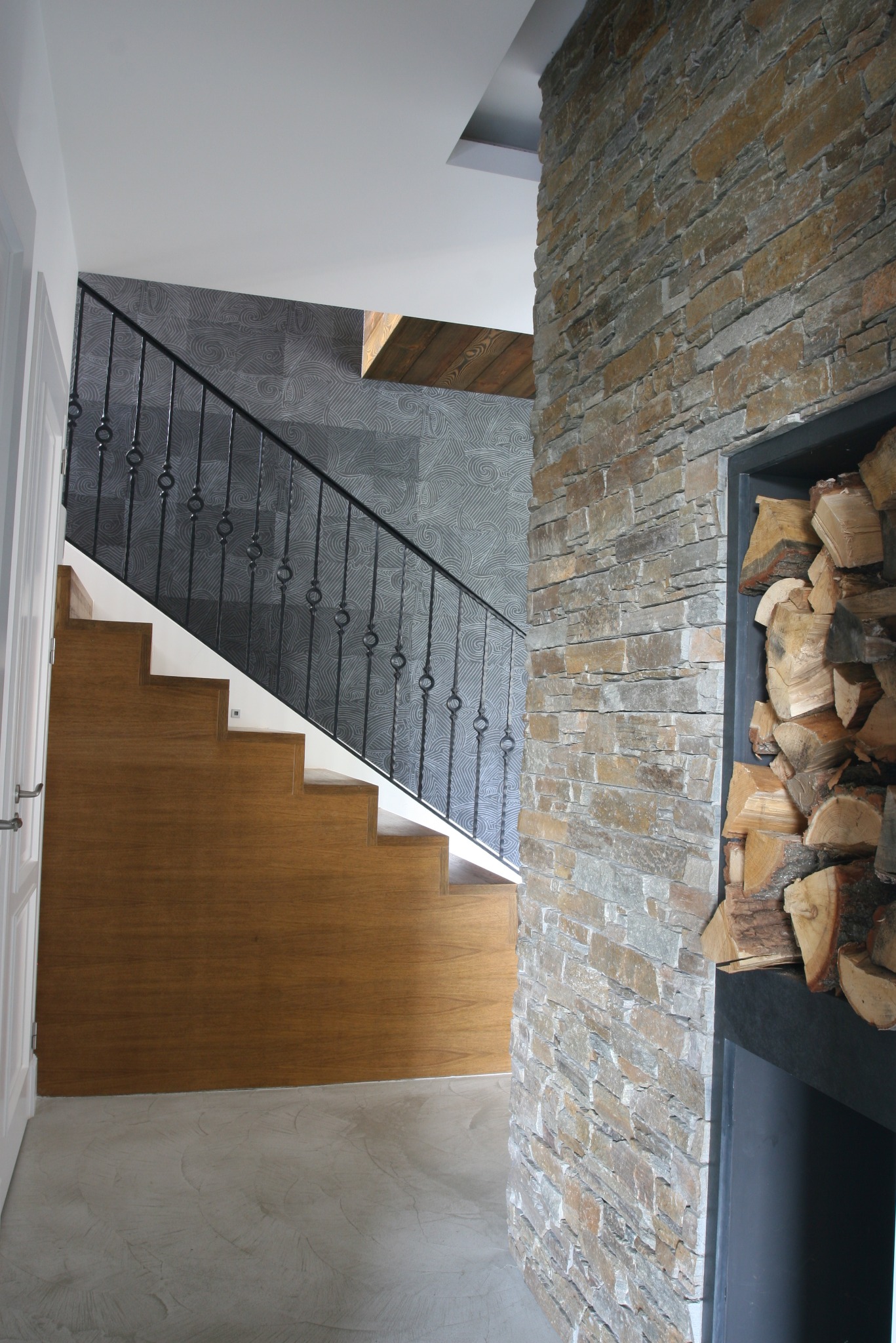 Drewniane schody z metalową balustradą na tle wzorzystej tapety, obok ściana z kamienia z wnęką na drewno kominkowe.