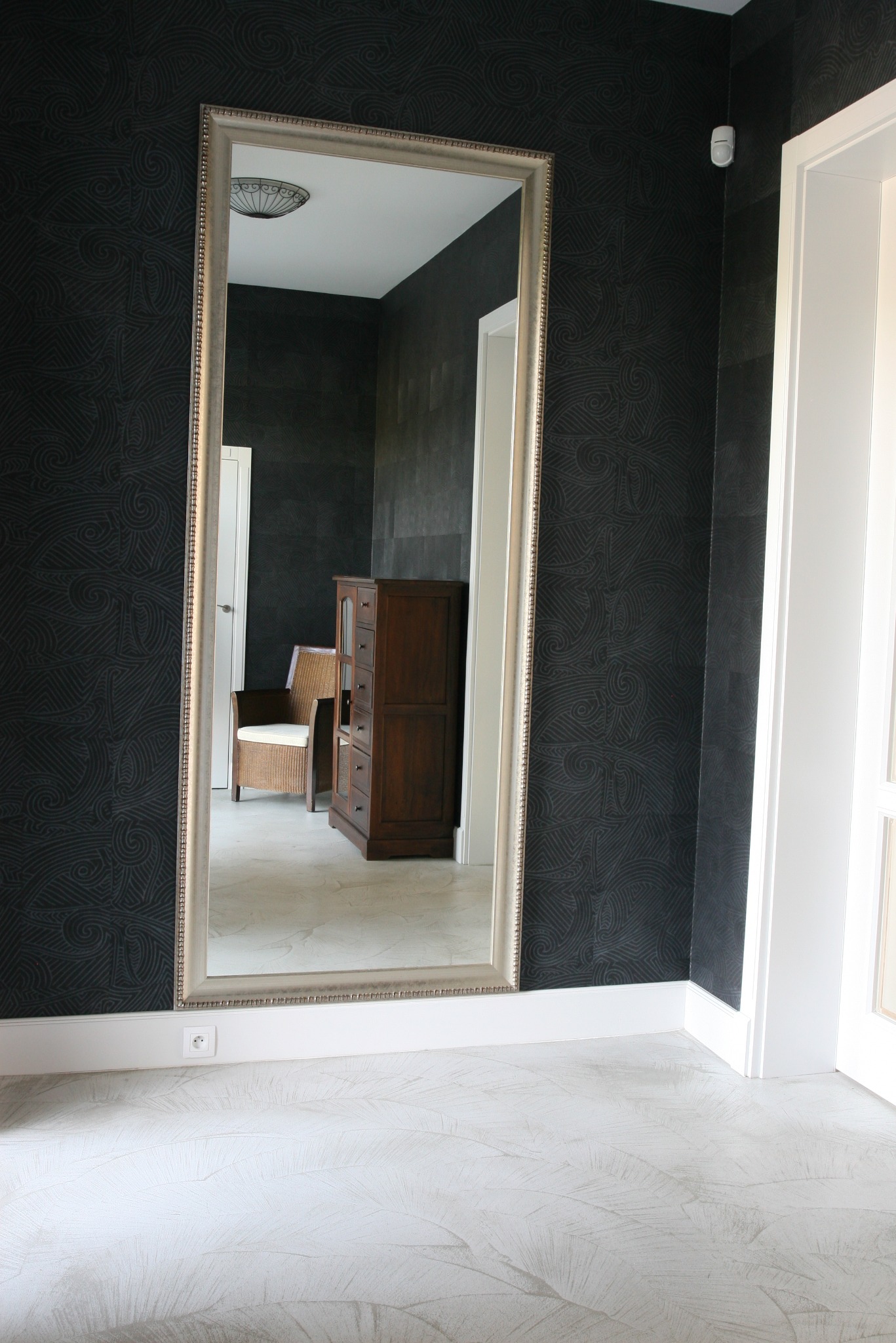 Wysokie lustro w srebrnej ramie odbija wnętrze pokoju z ciemną tapetą w geometryczne wzory, widoczna komoda i krzesło z wikliny.