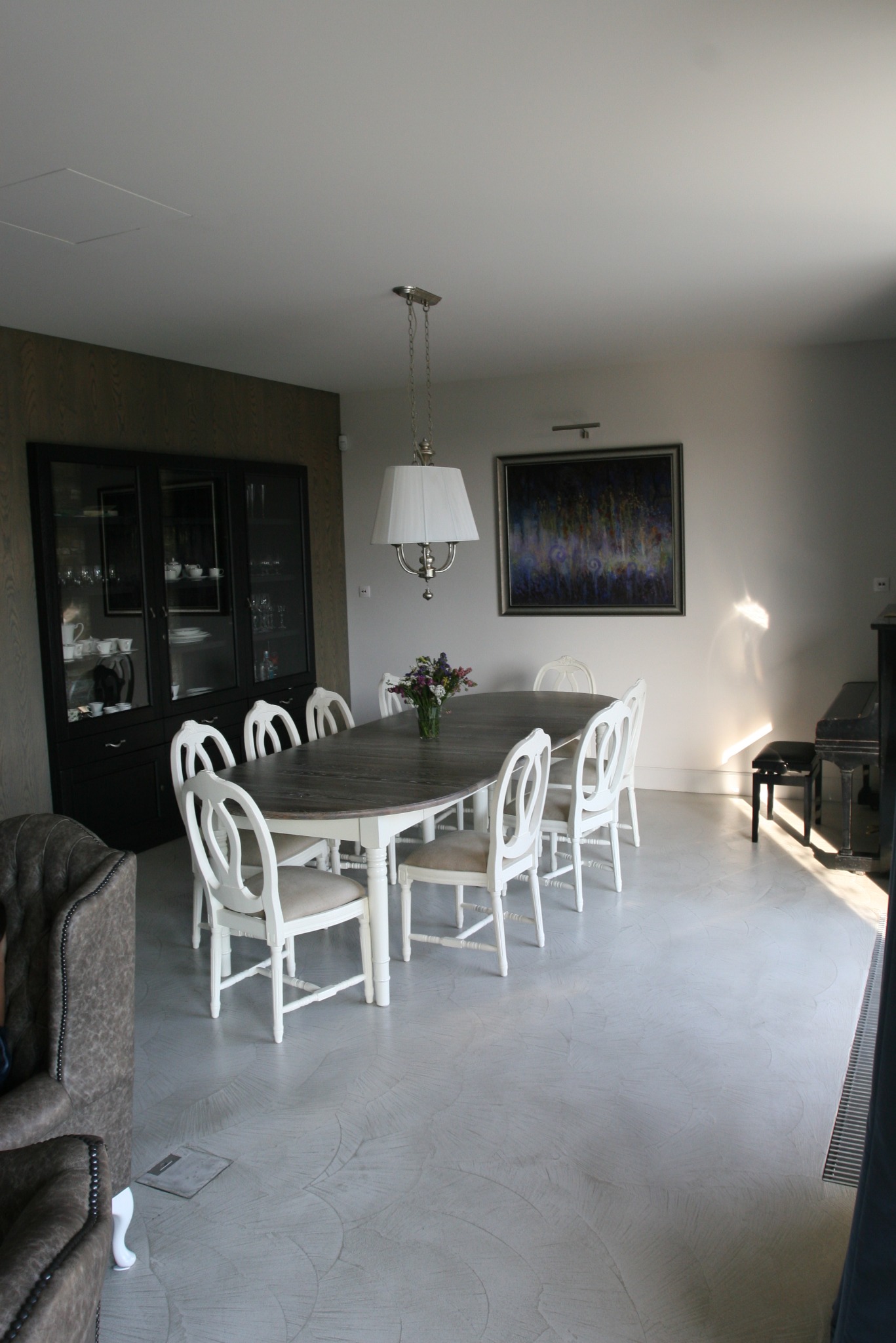 Jasne wnętrze jadalni z długim, drewnianym stołem otoczonym białymi krzesłami, pod wiszącą lampą z abażurem, na tle ciemnej witryny i obrazu na ścianie, z widocznym fortepianem w rogu.