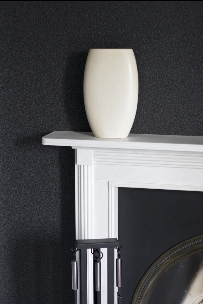 Kremowy wazon na białym kominku z narzędziami kominkowymi na tle ciemnej ściany.