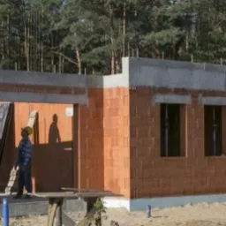 Budowa domu z czerwonej cegły ceramicznej, widoczni dwaj pracownicy w kaskach, jeden na dachu, drugi w otworze drzwiowym, w tle las.