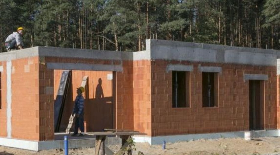 Budowa domu z czerwonej cegły ceramicznej, widoczni dwaj pracownicy w kaskach, jeden na dachu, drugi w otworze drzwiowym, w tle las.