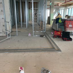 Firma Ogólnobudowlana Orzeł-Bud - Montaż metalowej konstrukcji ścian działowych w surowym stanie deweloperskim, widoczne profile, narzędzia w kufrach, przewody elektryczne i materiały budowlane, w tle okna i drabina