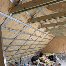 Firma Ogólnobudowlana Orzeł-Bud - Izolacja poddasza wełną mineralną, widoczne metalowe profile konstrukcyjne i drewniane belki, częściowo wykończona ściana z płyty OSB, bałagan na podłodze.