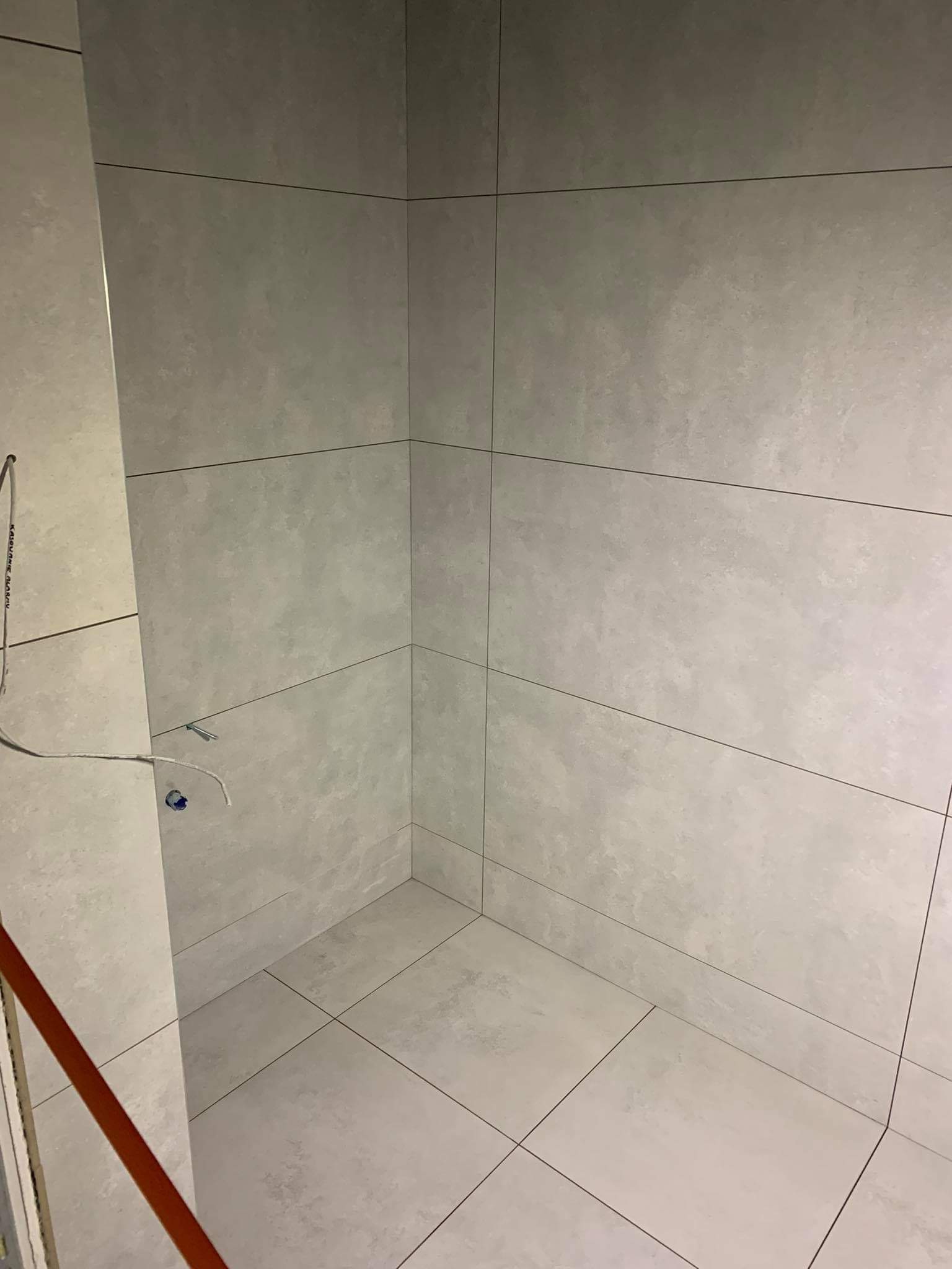 Świeżo ułożone, duże, szare płytki gresowe imitujące beton na ścianach i podłodze w rogu łazienki, widoczne przewody elektryczne i elementy instalacji wodnej przed armaturą.