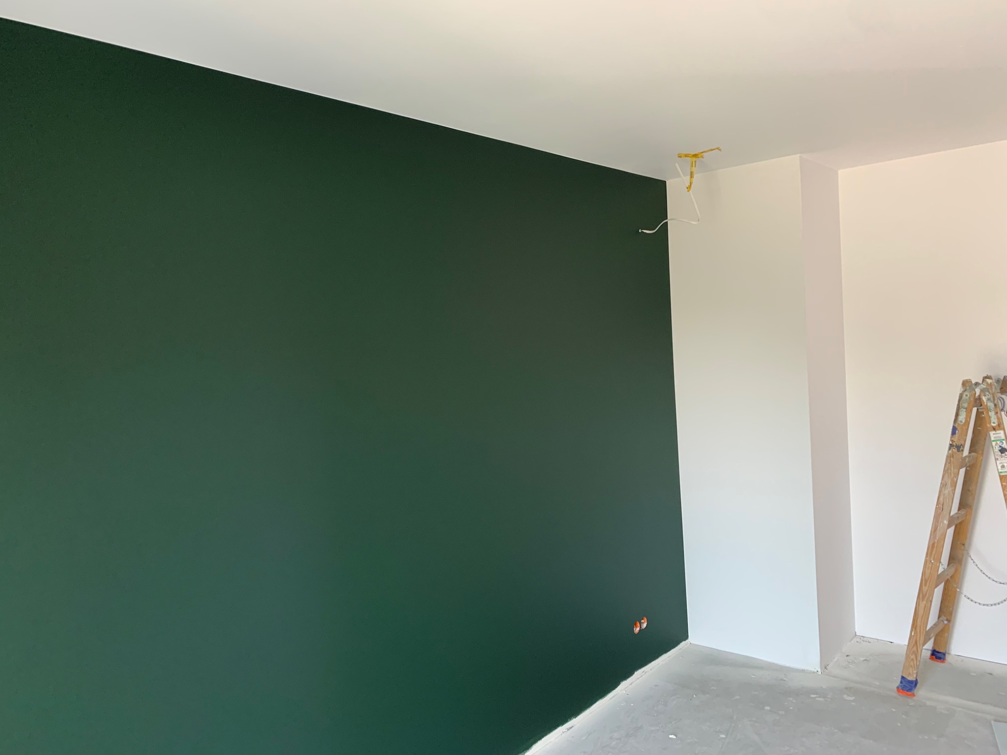 Świeżo pomalowana ściana na ciemnozielony kolor w narożniku pokoju, widoczne gniazdka elektryczne, biała ściana obok i drabina malarska z boku.