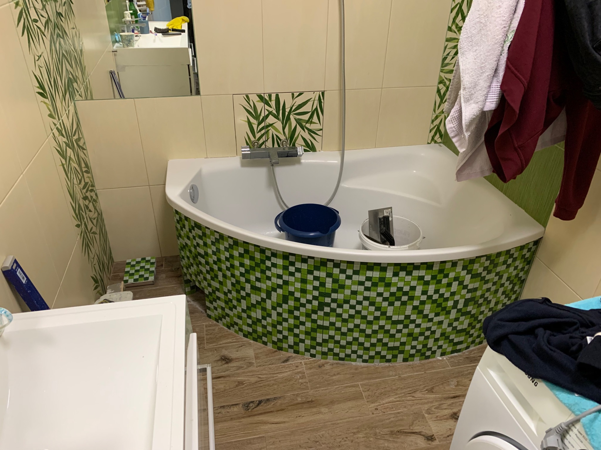 Biała wanna z obudową w zielono-białą szachownicę, niebieskie wiadro i narzędzia wewnątrz, płytki z motywem bambusa na ścianie, widok na fragment umywalki i pralki.