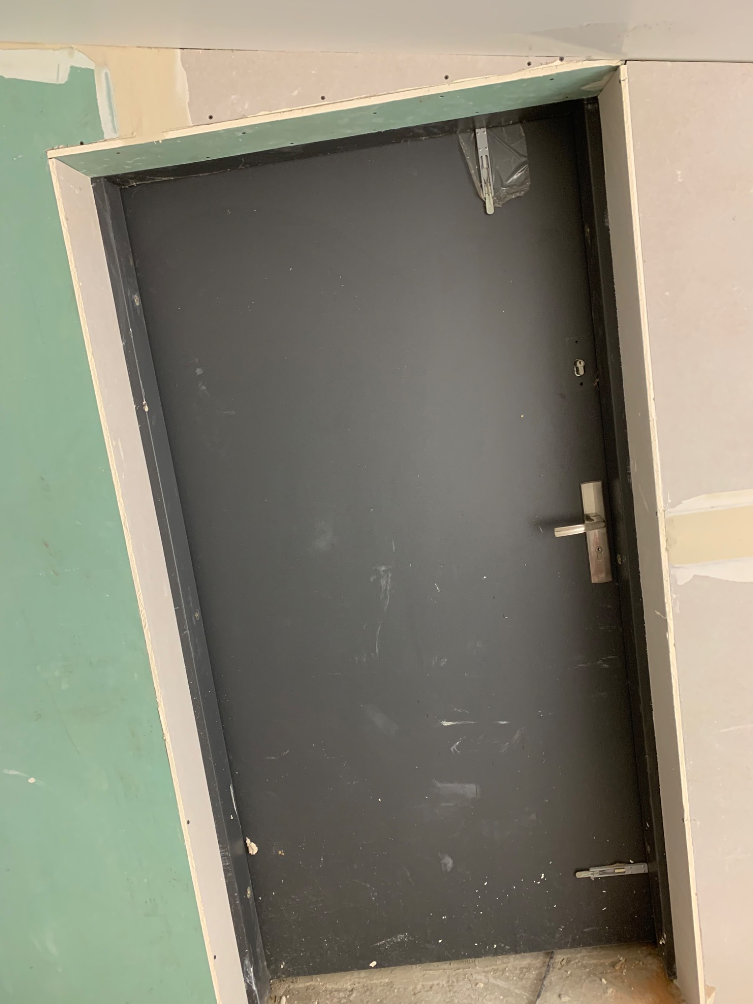 Ciemne drzwi z metalową klamką osadzone w ścianie z płyt gipsowo-kartonowych, widoczne ślady po szpachlowaniu i malowaniu.