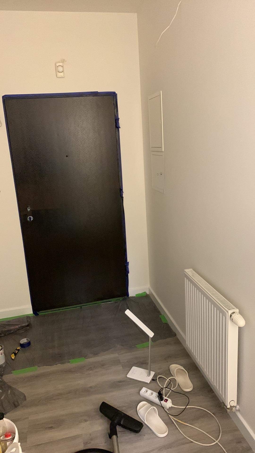 Przygotowanie do malowania: zabezpieczone drzwi taśmą malarską, folia ochronna na podłodze, grzejnik i ściany w jasnym odcieniu, lampka biurkowa, przedłużacz i kapcie na podłodze z paneli.