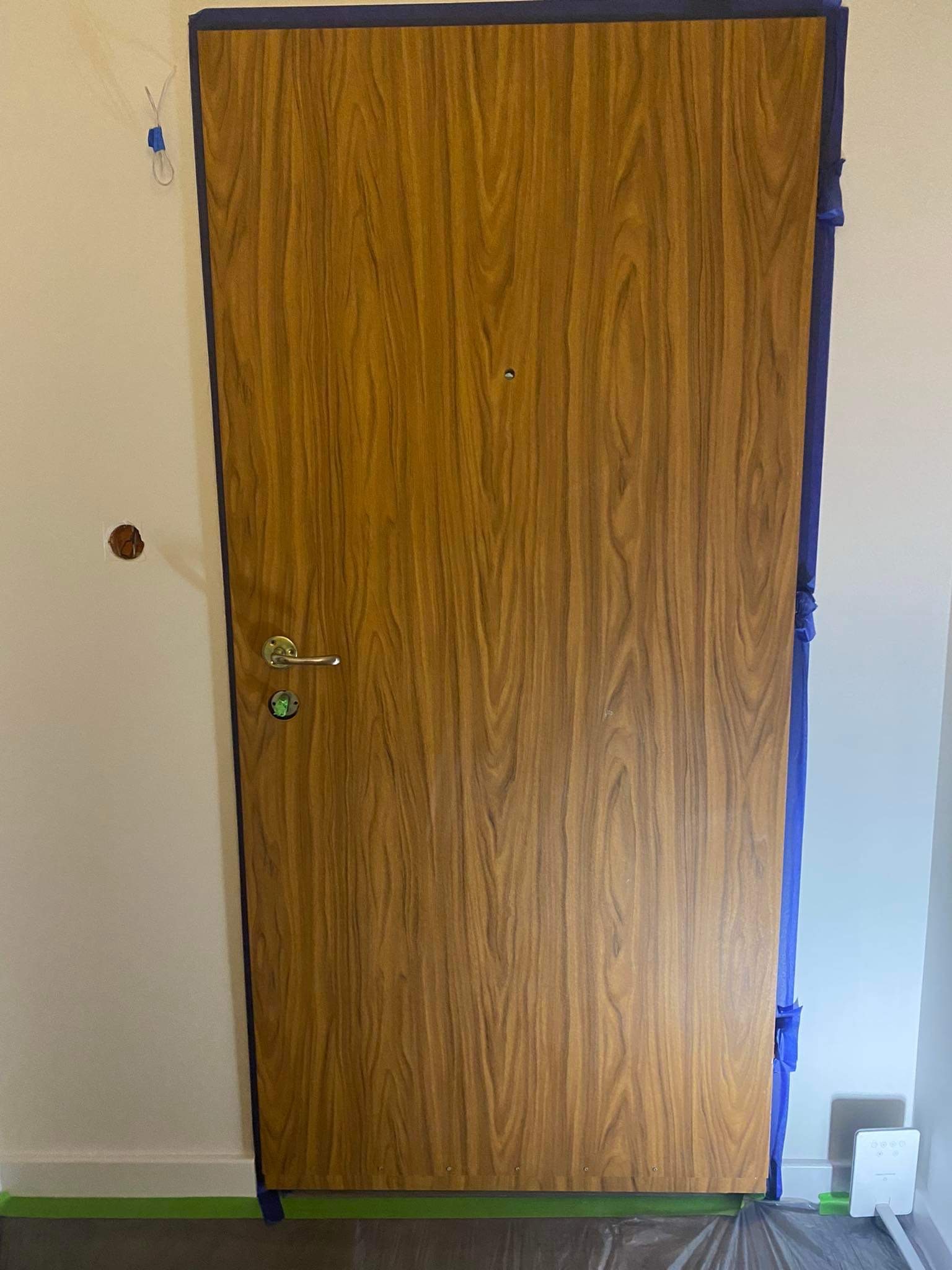Drzwi wejściowe z okleiną drewnopodobną, zabezpieczone niebieską taśmą malarską podczas prac wykończeniowych, widoczna dziura w ścianie po gniazdku elektrycznym.