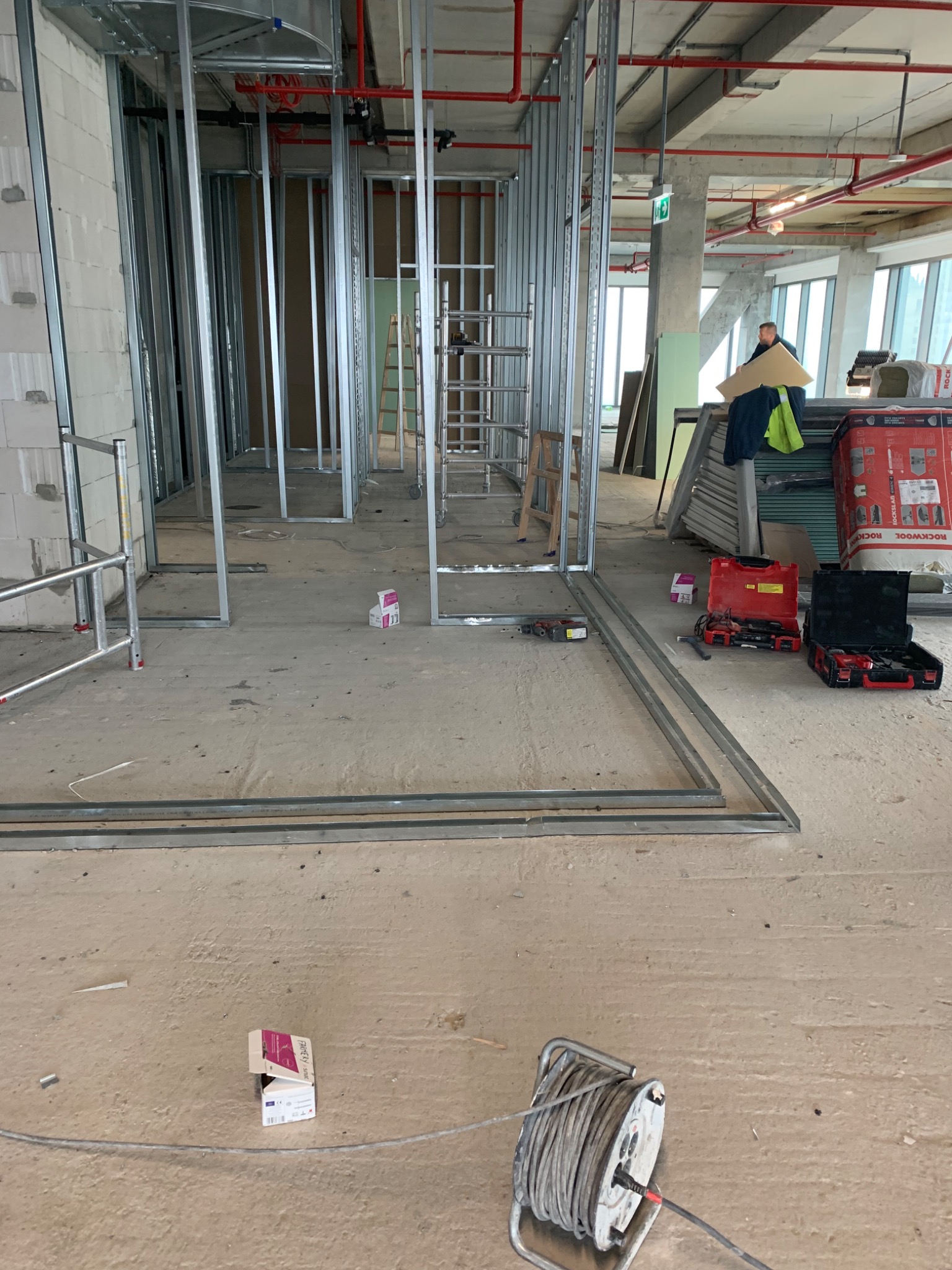 Montaż metalowej konstrukcji ścian działowych w surowym stanie deweloperskim, widoczne profile, narzędzia w kufrach, przewody elektryczne i materiały budowlane, w tle okna i drabina